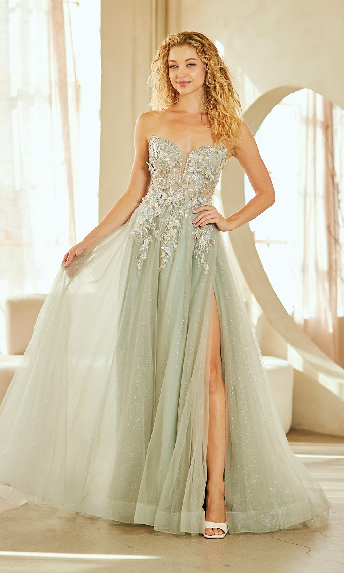 A woman stands indoors by large windows wearing the Strapless Glitter Prom Gown: Amelia Couture 4062 in sage, featuring a strapless design, A-line silhouette, 3-D floral appliqué, sheer bodice, and thigh-high slit.